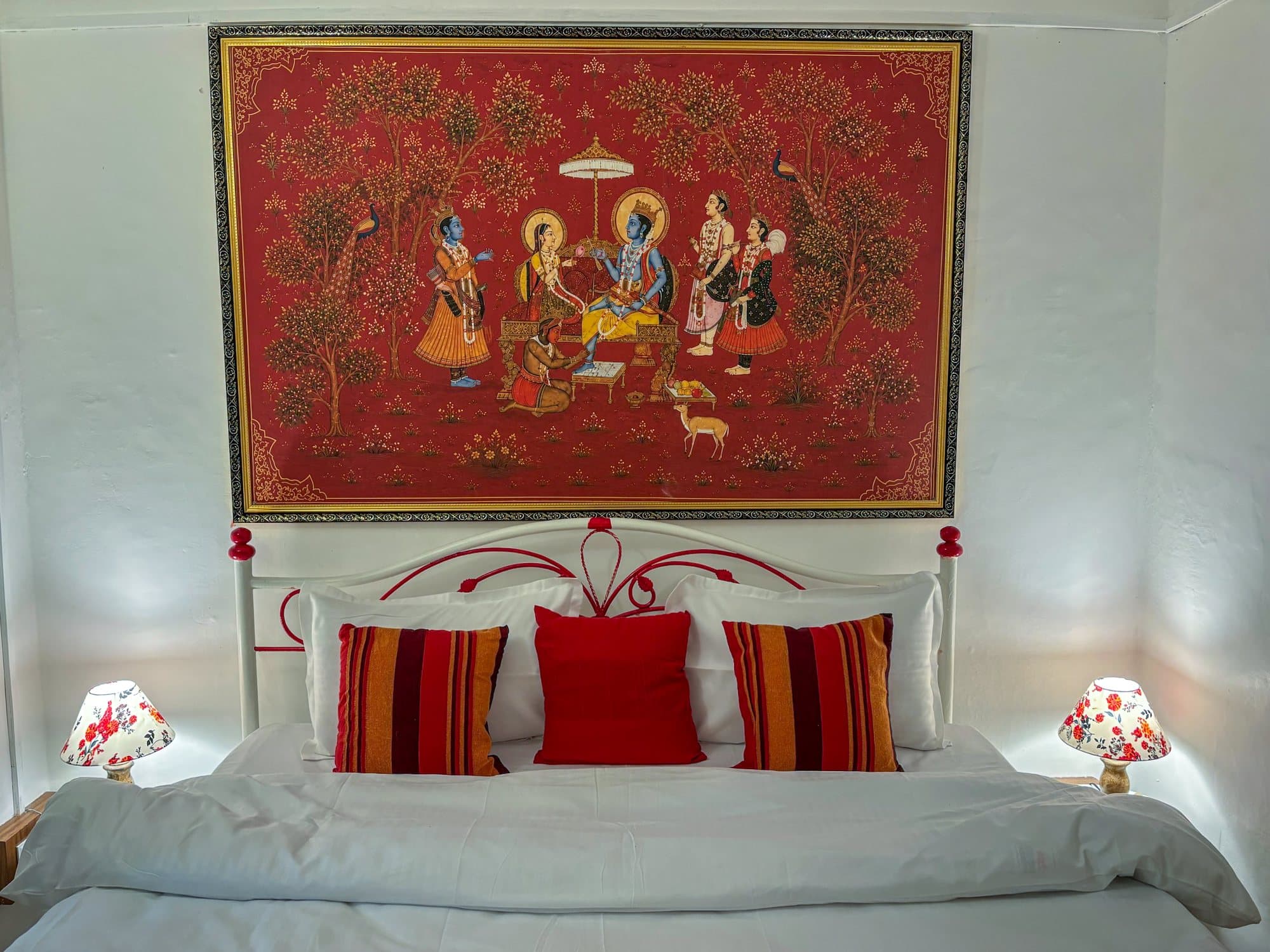 A bedroom at Shail Shikhar with a red Pichwai above a white iron bed
